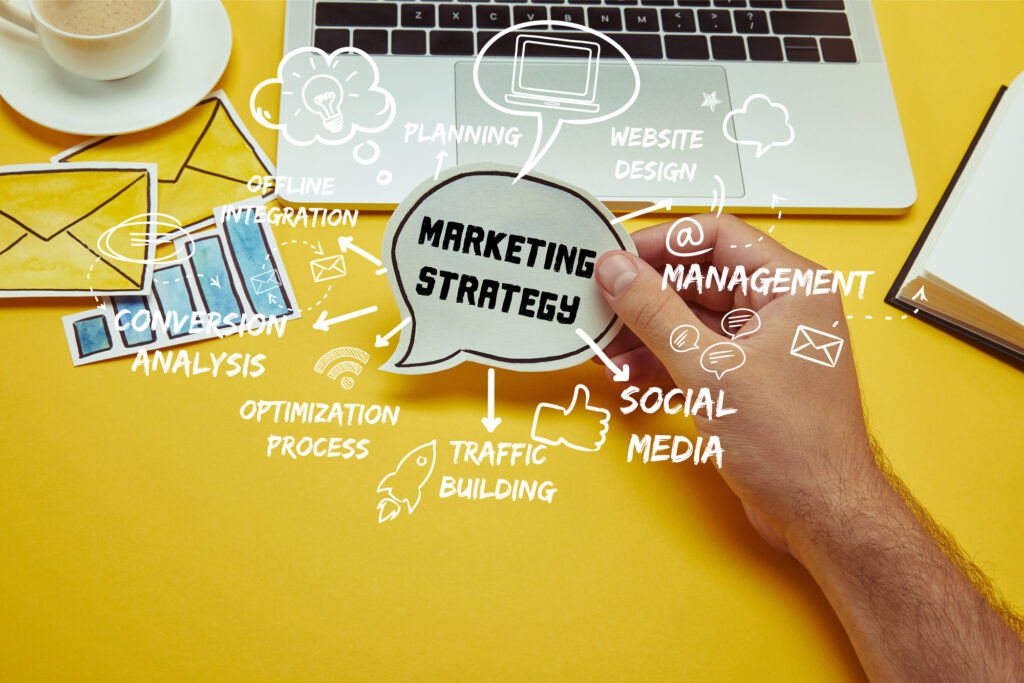 A person holding up a cutout with the bubble that says "Marketing Strategy." Surrounded by it includes the words Planning, Website Design, Management, Social Media, Traffic Building, Optimization Process, Conversion Analysis, and Offline Integration.
